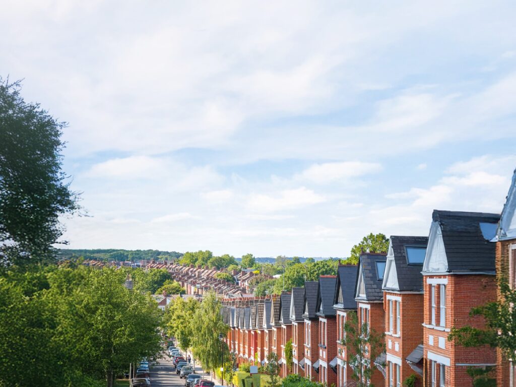 A hillside street of red-brick Victorian terraced houses in bright daylight, looking down the road past parked cars towards rooftops in the distance